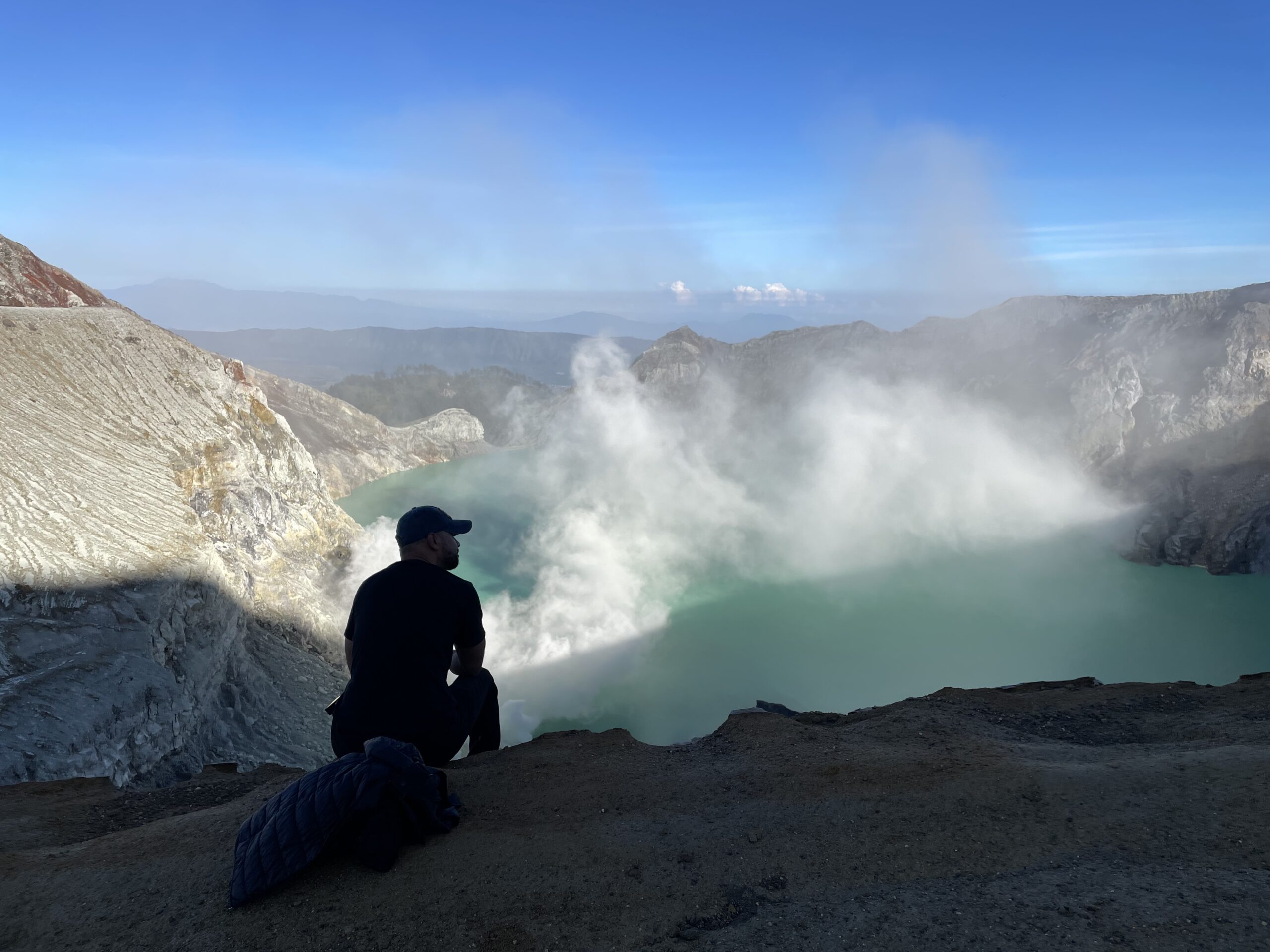 The turquoise acid lake of Kawah Ijen at sunrise, smoke rising, a lone figure silhouetted at the crater rim